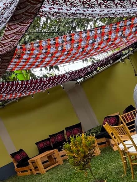 Kitenge cloth strung overhead as a canopy above pallet benches with patterned cushions, beside the lawn