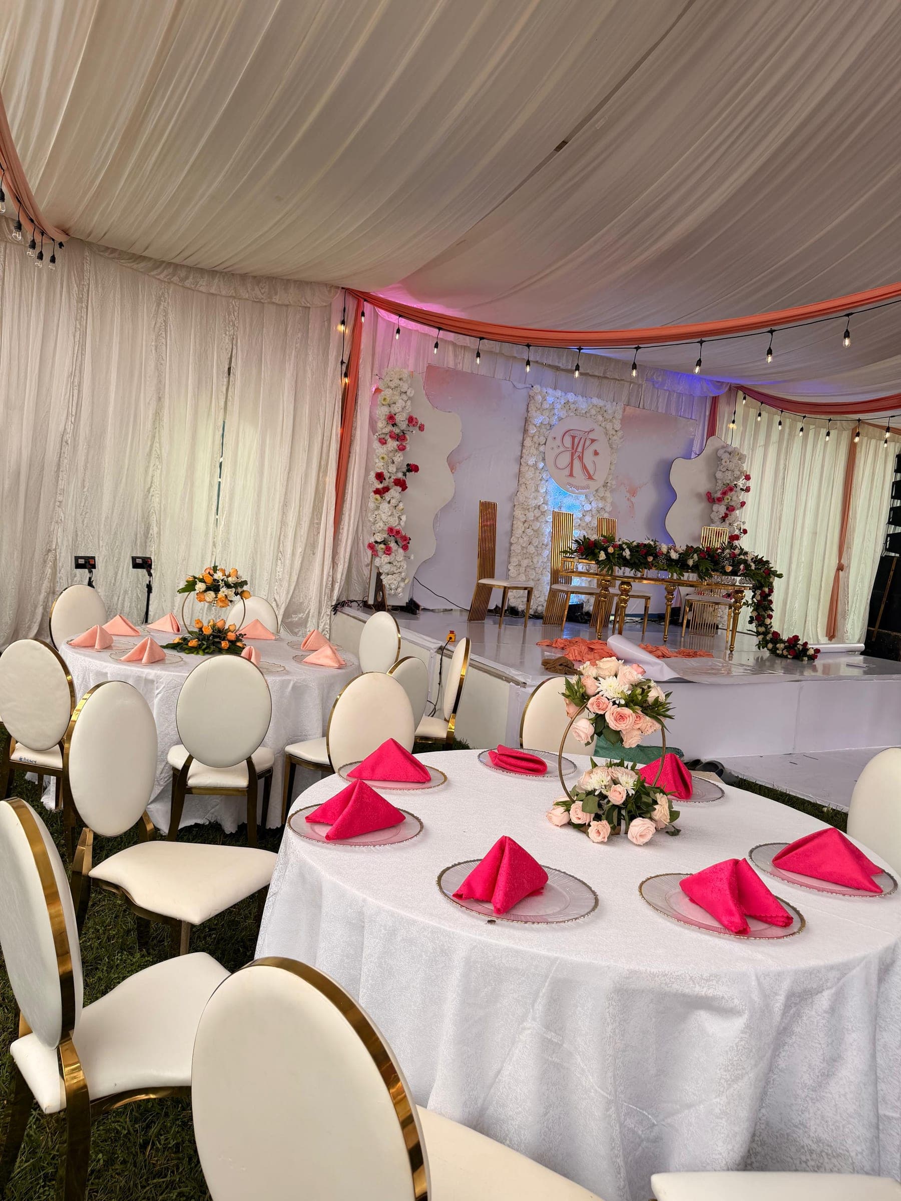 Round tables set with fuchsia and peach napkins in front of a monogrammed flower wall, under coral drapes and festoon lighting